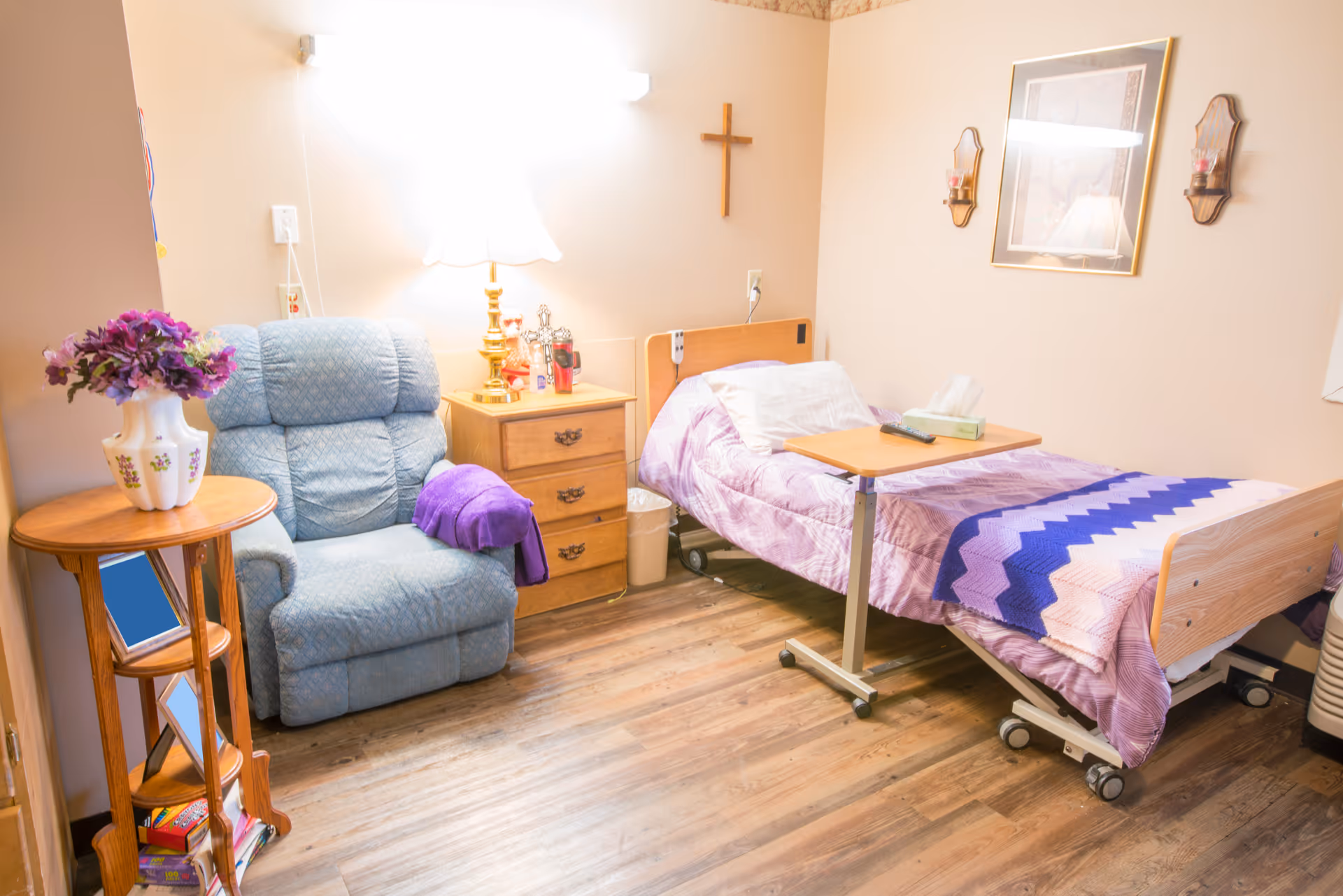 A cozy senior care room with a single hospital-style bed covered in a purple and pink blanket, a wooden bedside table with a lamp, a blue recliner chair with a purple throw, a small wooden side table with a vase of purple flowers, and wall decorations including a cross and framed artwork.