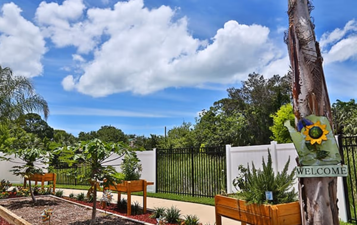 Raised garden beds and plants beside a fenced walkway with a tree displaying a 'WELCOME' sign under a blue sky.