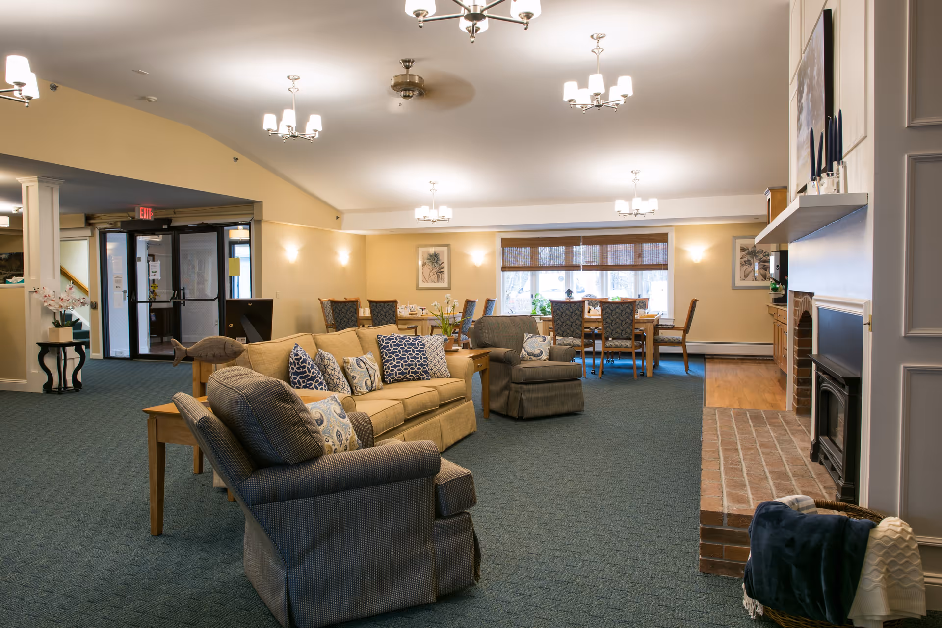 A spacious and well-lit common area in a senior living facility featuring comfortable armchairs and a sofa with patterned cushions arranged around a brick fireplace. In the background, there is a dining area with a wooden table and chairs near large windows with blinds. The room has carpeted flooring, warm yellow walls, and multiple ceiling light fixtures.