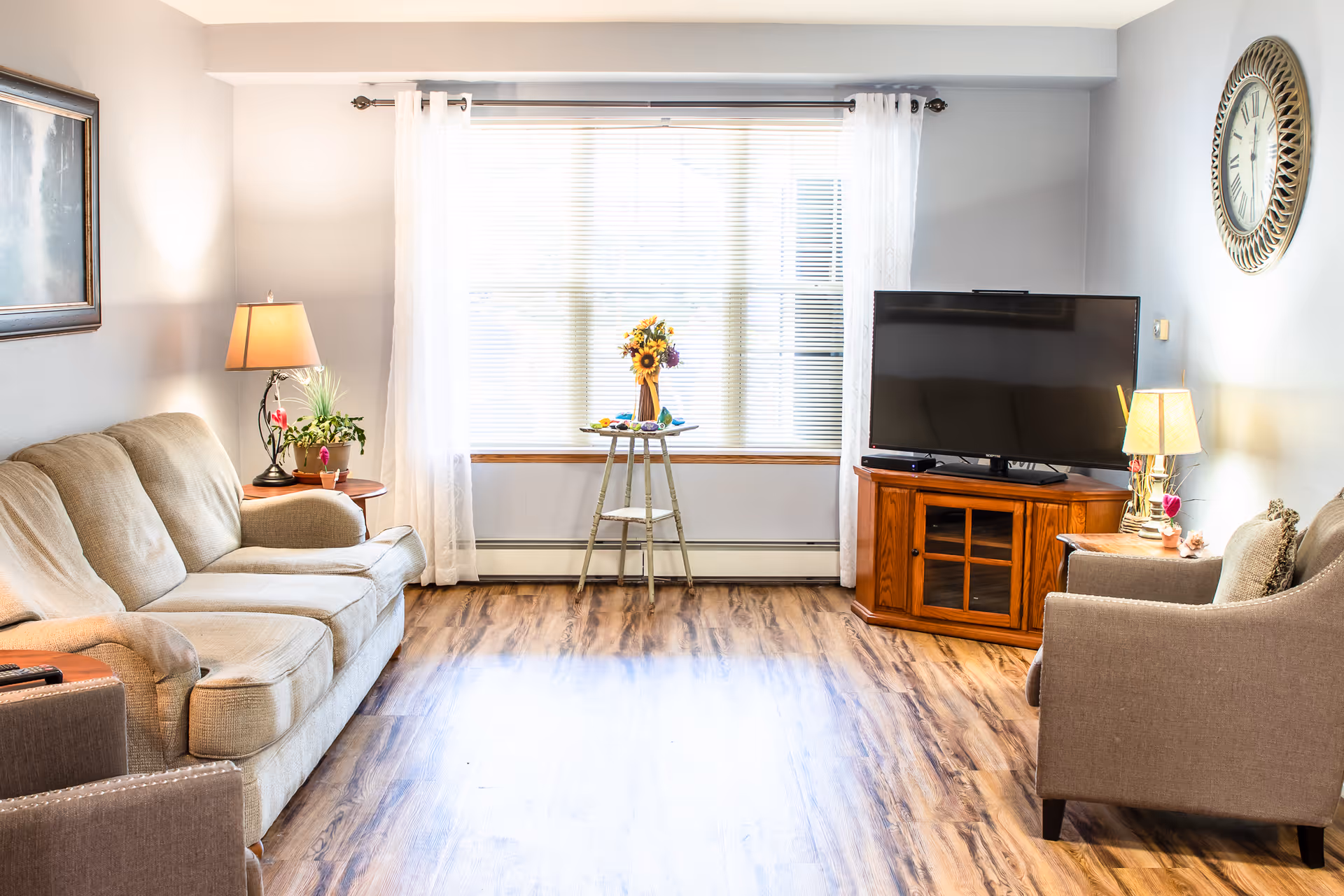 Bright living room with a sofa and armchair facing a TV on a wooden stand, a large window with sheer curtains, and wood-look flooring.