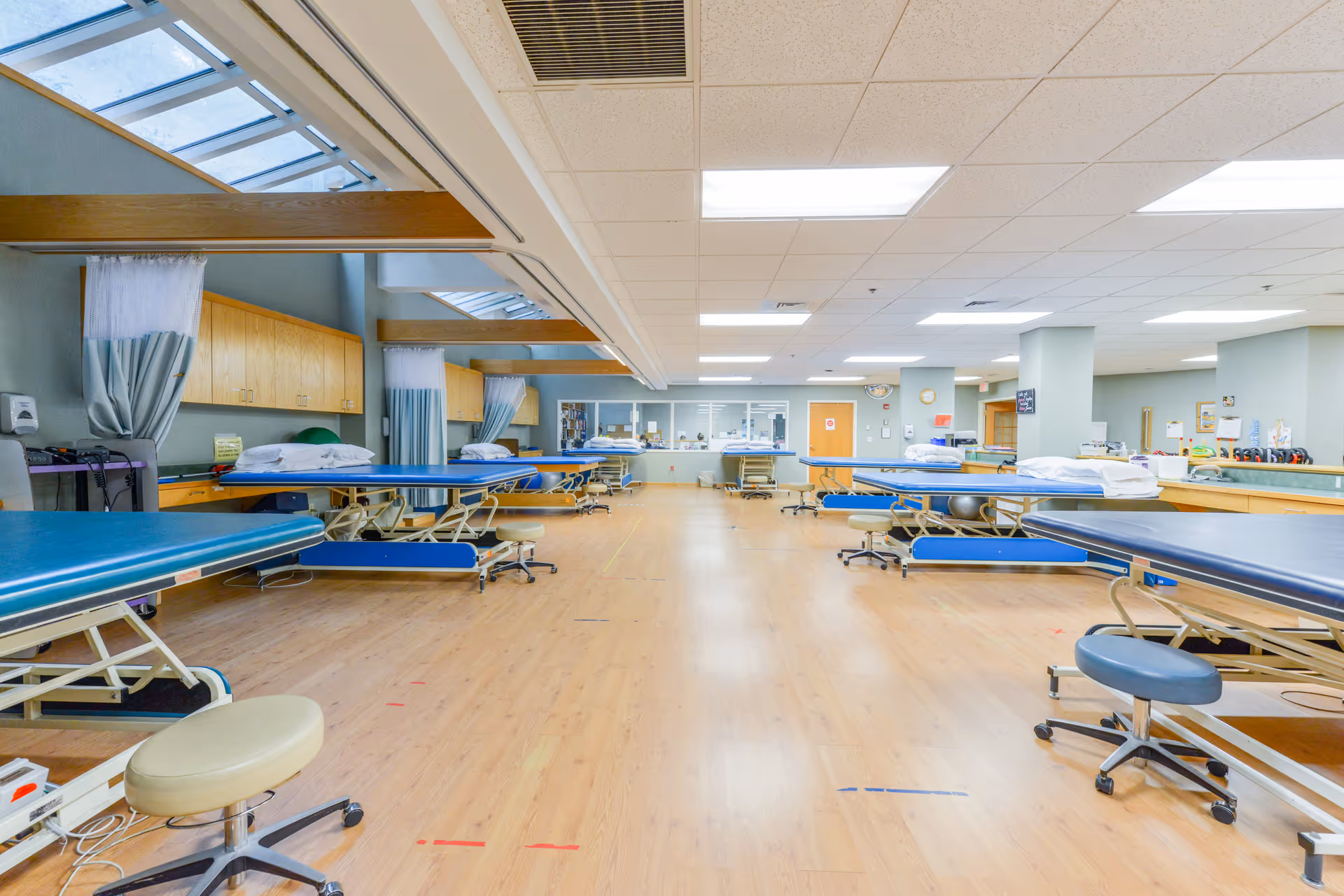 Large, bright rehabilitation/therapy room with multiple treatment tables, stools, and exercise equipment.
