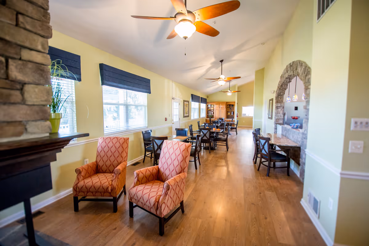 A spacious, well-lit common area with wooden floors, two patterned armchairs near a stone fireplace, multiple tables and chairs arranged along the wall with windows, ceiling fans with lights, and a stone archway leading to another room.