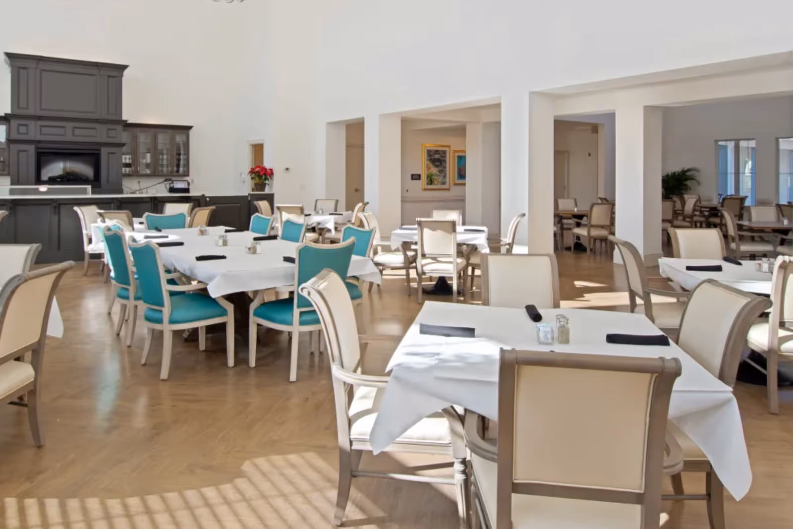 Bright dining room with multiple tables set with white tablecloths and teal and beige chairs.