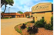 Exterior view of LakePoint El Dorado, a single-story building with a brown roof and brick walls, surrounded by a paved driveway and landscaped garden beds with shrubs and small plants under a partly cloudy sky.