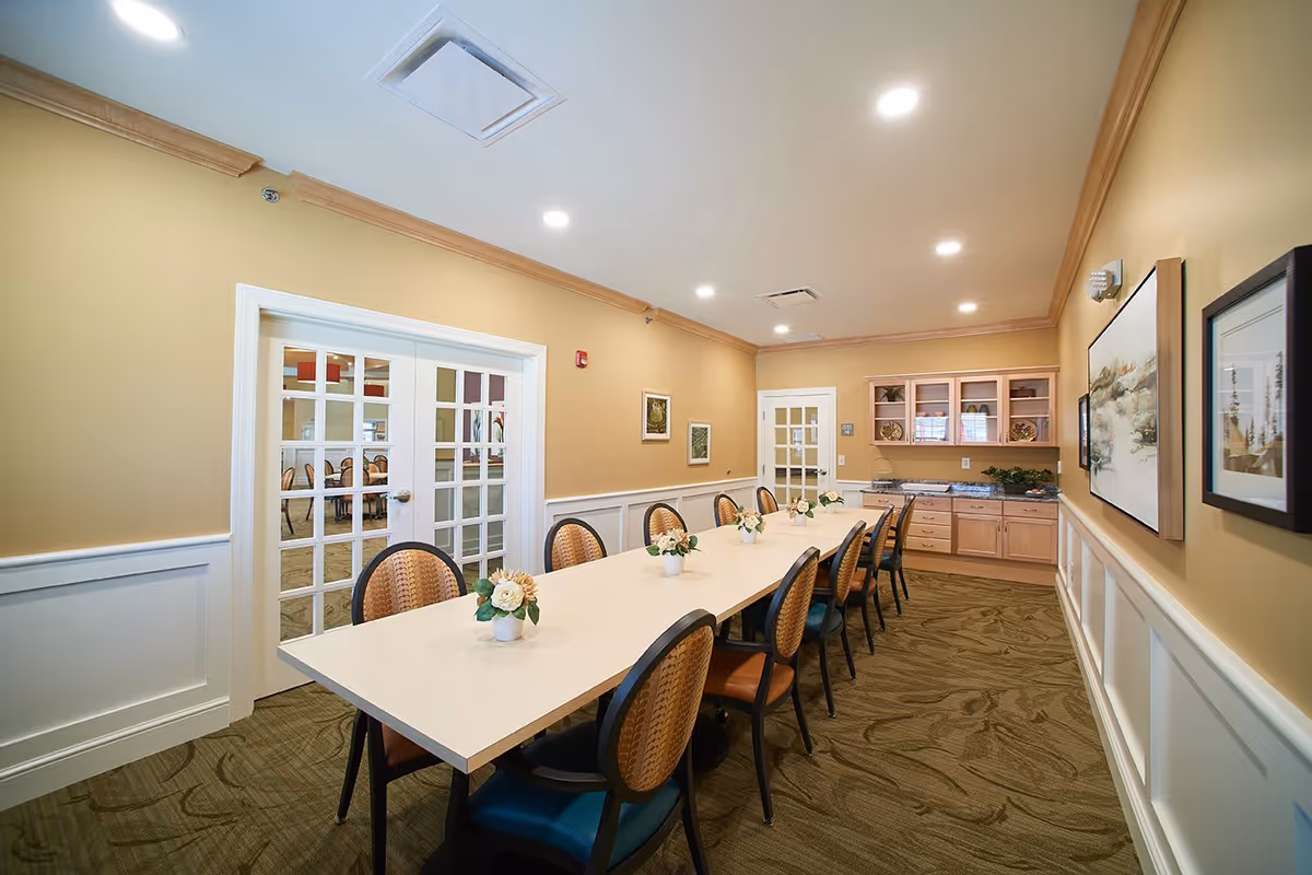 A long dining table with ten chairs arranged around it in a well-lit room with beige walls and carpeted floor. The table has small flower arrangements placed along its center. There are glass-paneled double doors on one side and a cabinet with countertop and decorative items on the far end. Framed artwork is hung on the walls.