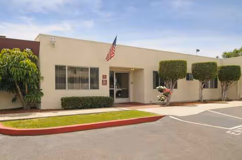 Single-story beige building with an American flag, entrance, windows, and manicured shrubs.