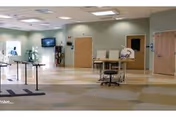 Spacious interior rehab/therapy room with parallel walking bars, chairs, a wall-mounted TV, and open tiled floor space.