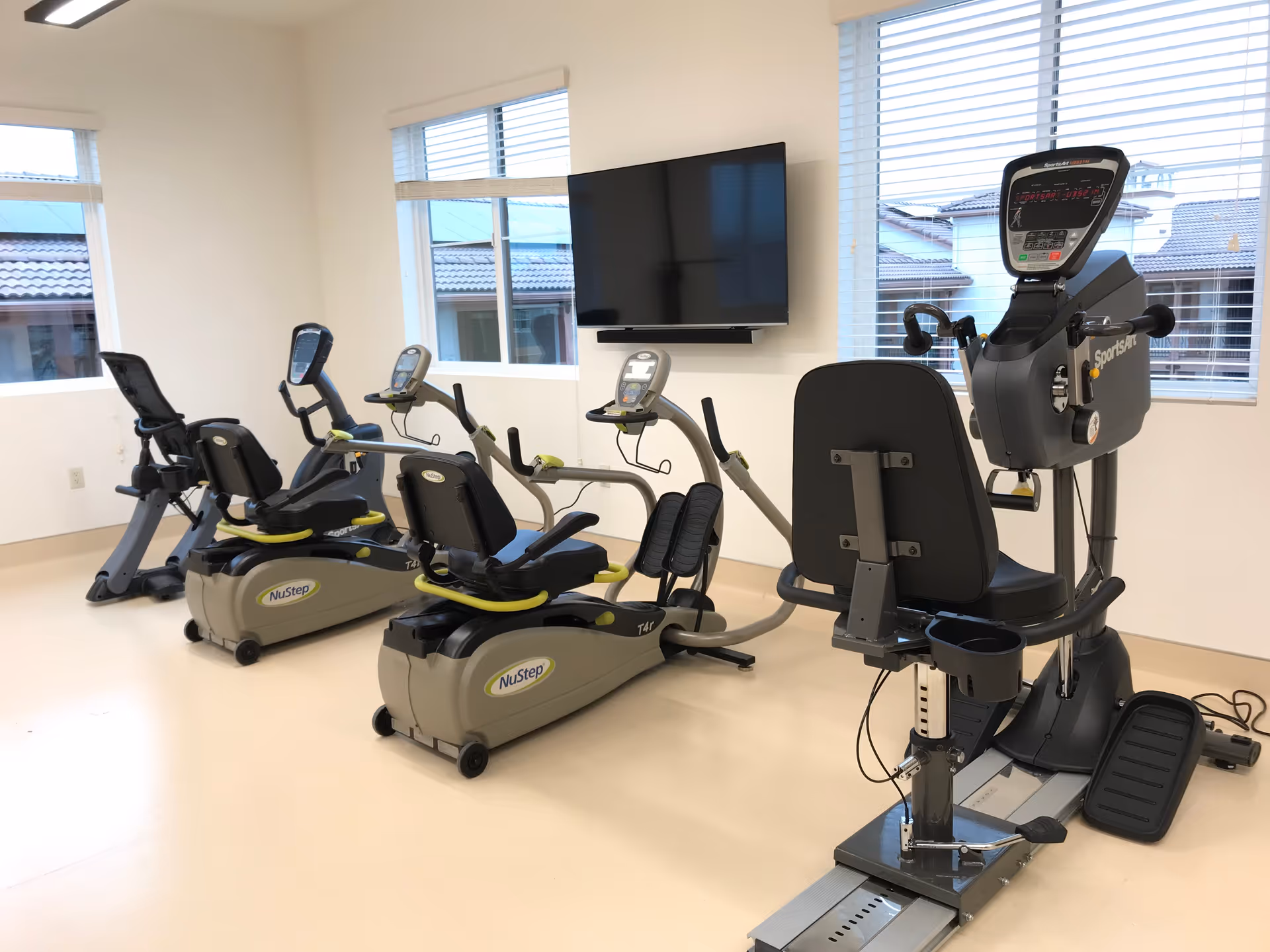 Bright interior fitness room with several recumbent exercise bikes facing a wall-mounted TV and windows.