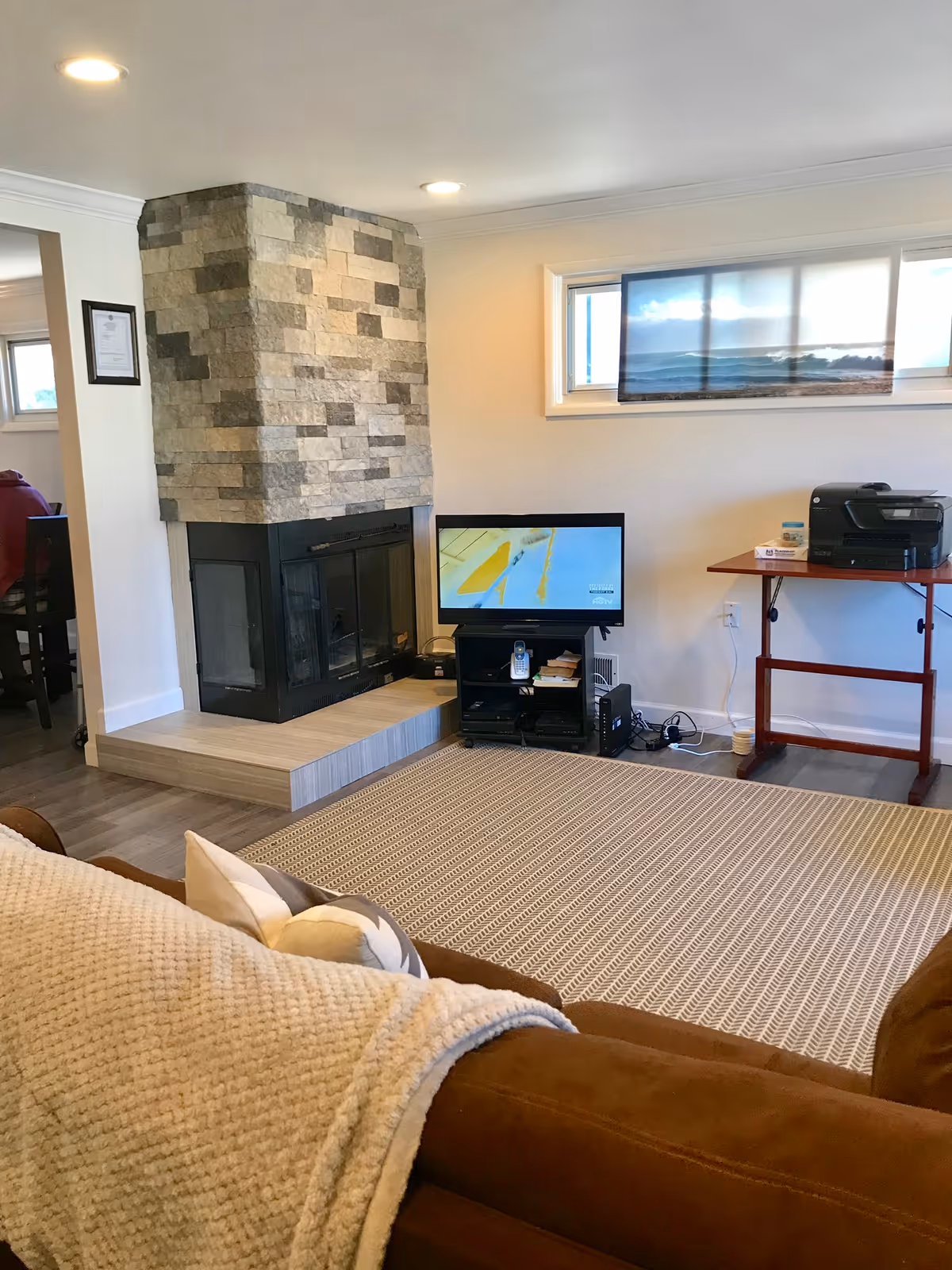 Cozy living room with a stone fireplace, a small TV on a stand, a brown couch with a beige throw blanket and pillows, a patterned area rug, and a wooden table with a printer on it. There is a window above the TV letting in natural light.