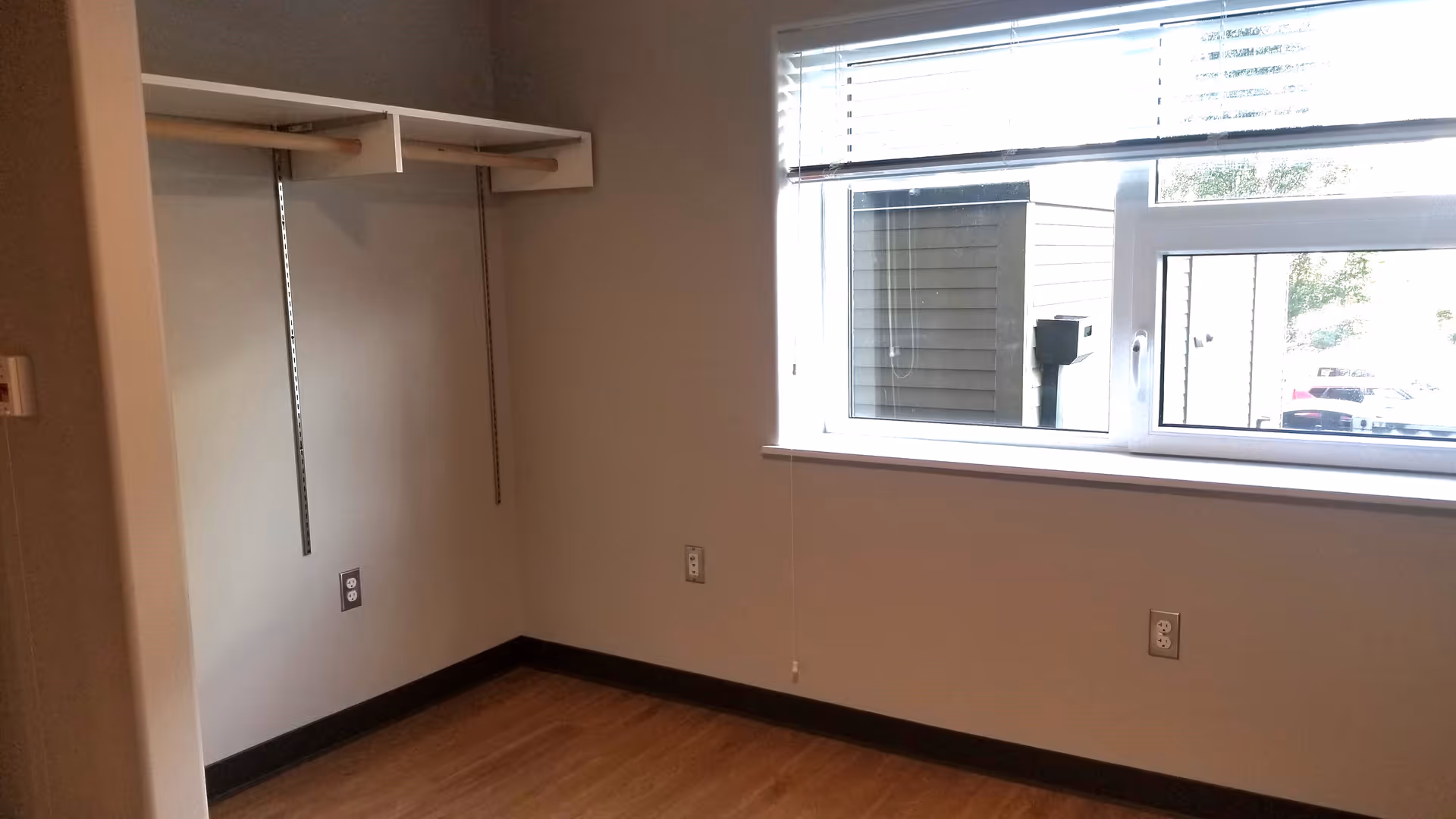 Empty small room with wood flooring, open closet shelving on the left and a large window with blinds on the right.