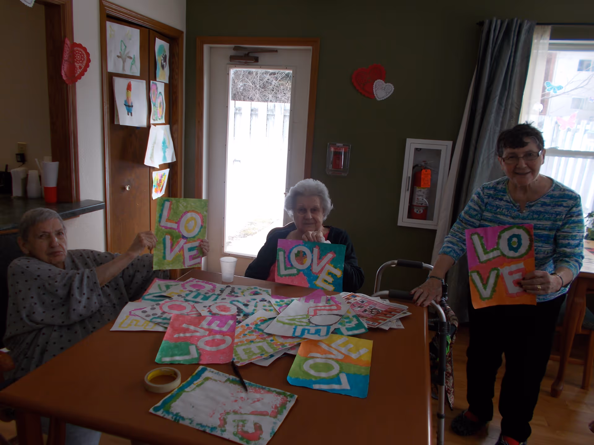 Three elderly women in a room holding colorful artwork with the word 'LOVE' painted on them. They are seated and standing around a table covered with similar paintings and art supplies. The room has green walls, a door with a window, and children's drawings taped to a wooden door.