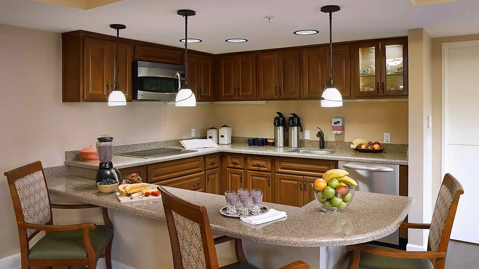 A kitchen area with wooden cabinets, granite countertops, and three pendant lights hanging from the ceiling. There are two wooden chairs with patterned cushions at the counter. On the counter, there is a blender with a purple smoothie, a bowl of blueberries, a cutting board with bananas and strawberries, a tray with four glasses of purple smoothie, and a bowl of assorted fruits including bananas, apples, and oranges. The kitchen also features a microwave, a dishwasher, and two coffee dispensers.