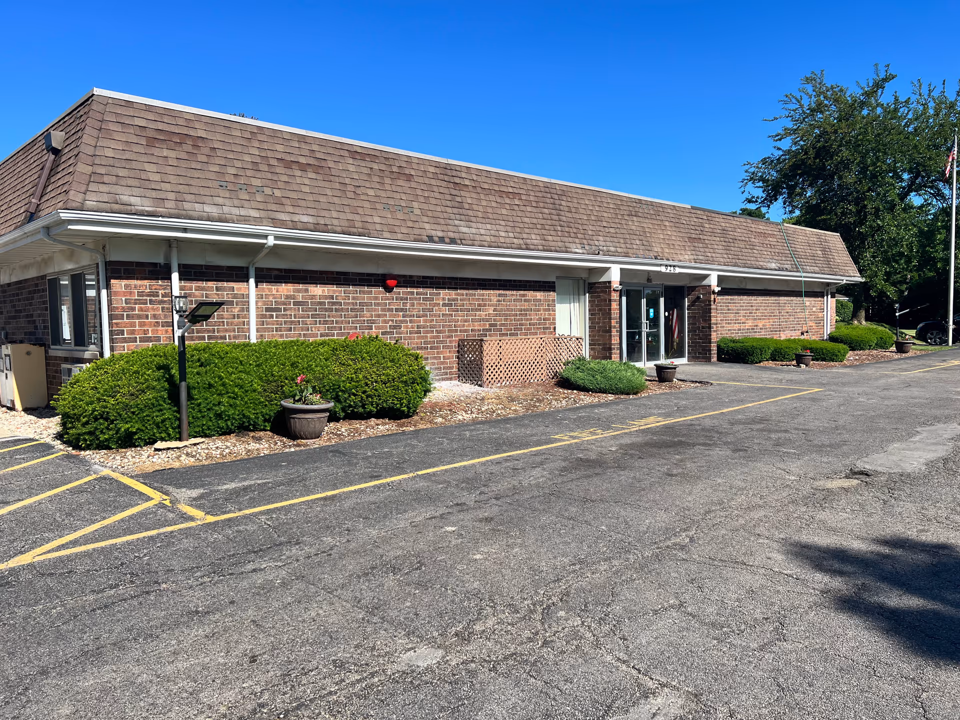 Exterior view of a single-story brick building with a brown shingled roof, surrounded by small bushes and potted plants. The building has a glass entrance door and is situated next to a paved parking area with yellow painted lines. The sky is clear and blue.