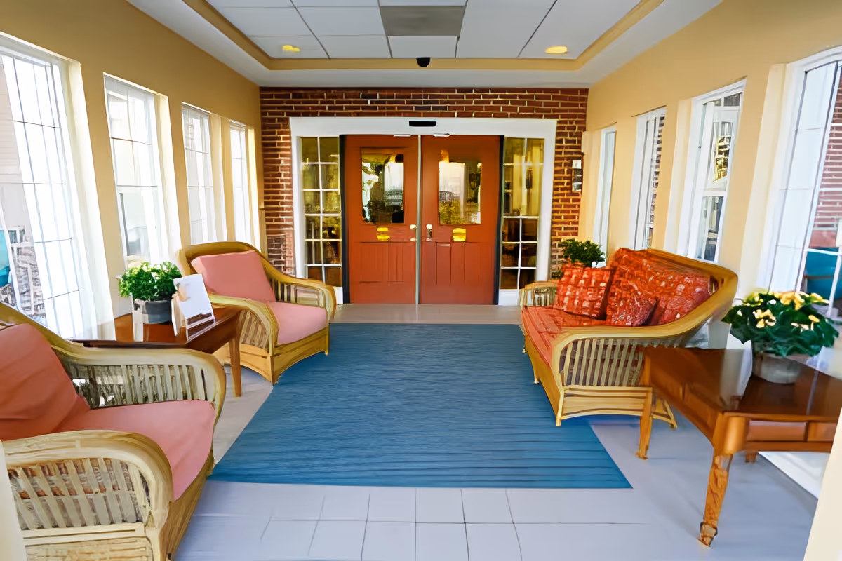 A bright and inviting seating area with wicker furniture including two armchairs with pink cushions and a sofa with red patterned cushions. The space has large windows on both sides letting in natural light, a blue rug on the tiled floor, potted plants on side tables, and double wooden doors with glass panels at the far end.