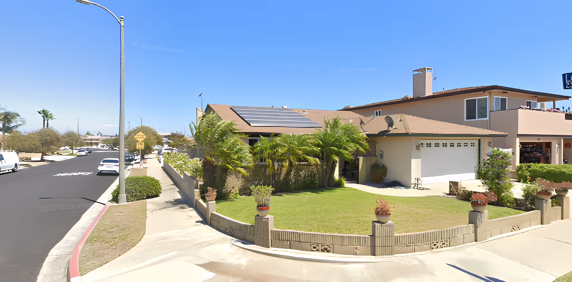 Exterior view of a residential building with a well-maintained lawn, palm trees, and a driveway leading to a garage. The building has solar panels on the roof and is situated on a corner lot with a sidewalk and street visible.