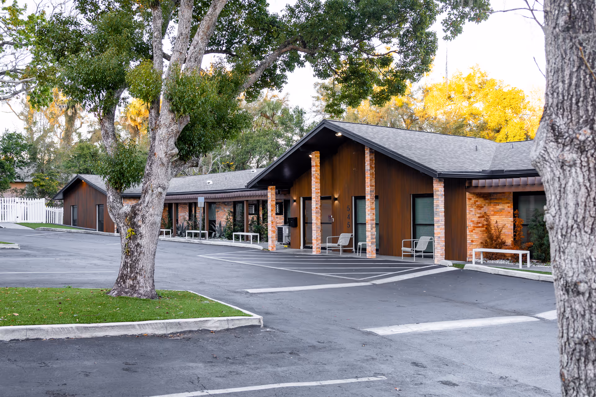 Exterior view of a single-story building with a sloped roof, brick and wood paneling, and several windows. The building is surrounded by trees and has a paved parking area with marked parking spaces in front. There are benches placed near the entrance and along the building.