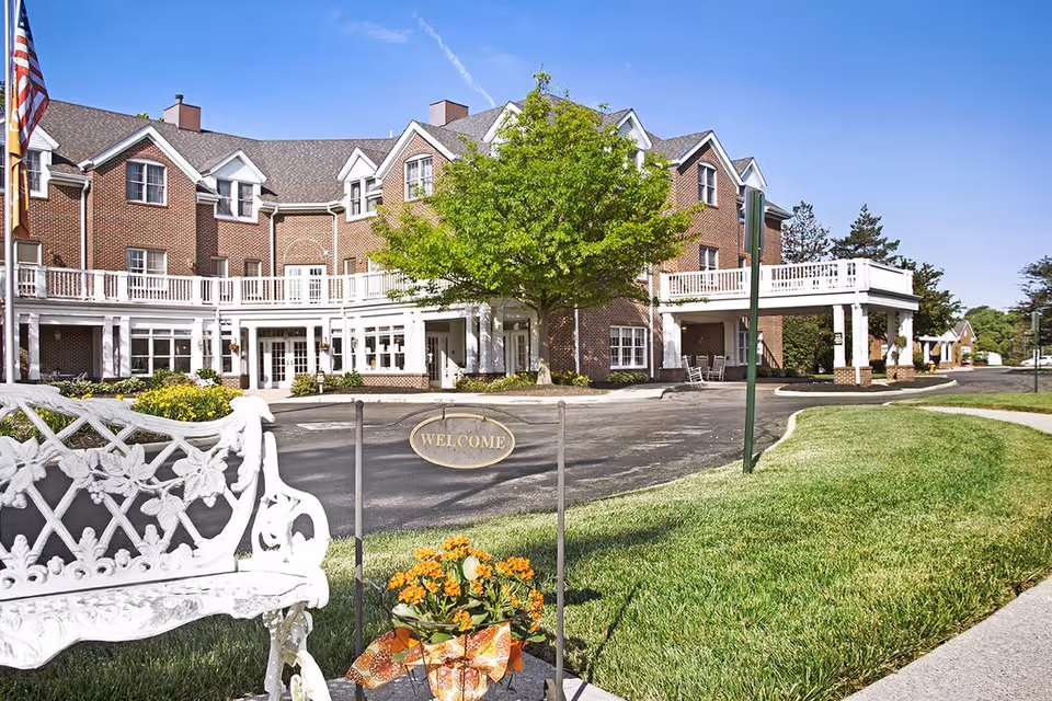 Front exterior of a brick senior living building with a white decorative bench, a small 'WELCOME' sign, flowers, and a landscaped driveway.