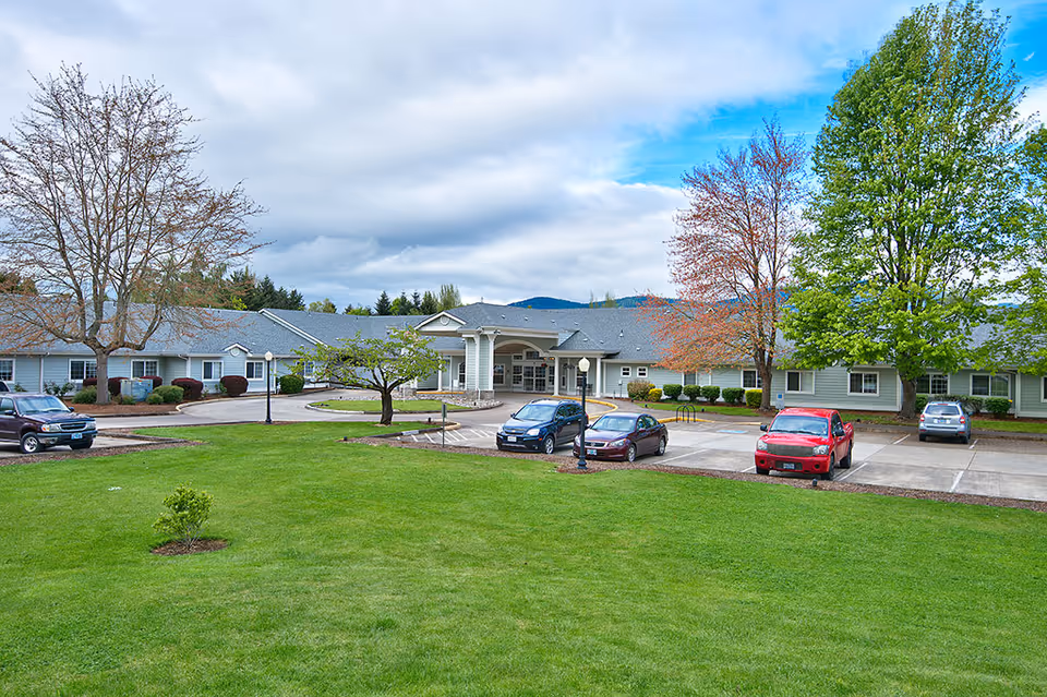 Exterior view of Spring Valley Assisted Living facility showing a single-story building with a covered entrance, surrounded by a well-maintained lawn, several trees, and a parking lot with a few parked cars under a partly cloudy sky.