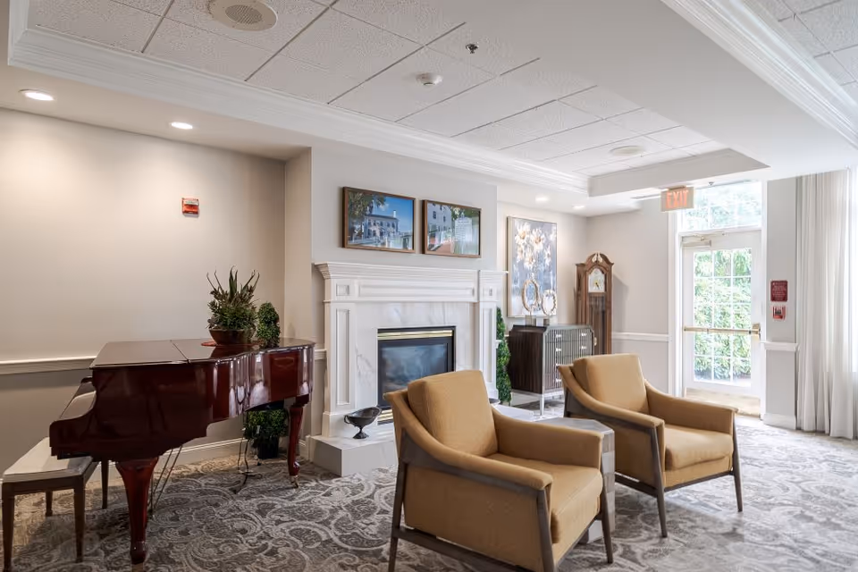 A bright and elegant common area featuring a polished grand piano with a bench, two beige armchairs, a white fireplace with framed pictures above it, a grandfather clock, and a cabinet. The room has a patterned carpet, light-colored walls, and large windows letting in natural light.