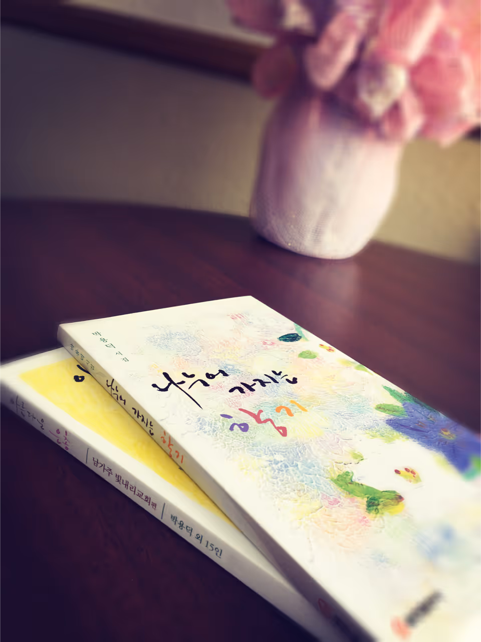 Two books with colorful covers featuring Korean text are placed on a dark wooden table. In the background, there is a blurred pink flower arrangement in a white vase.