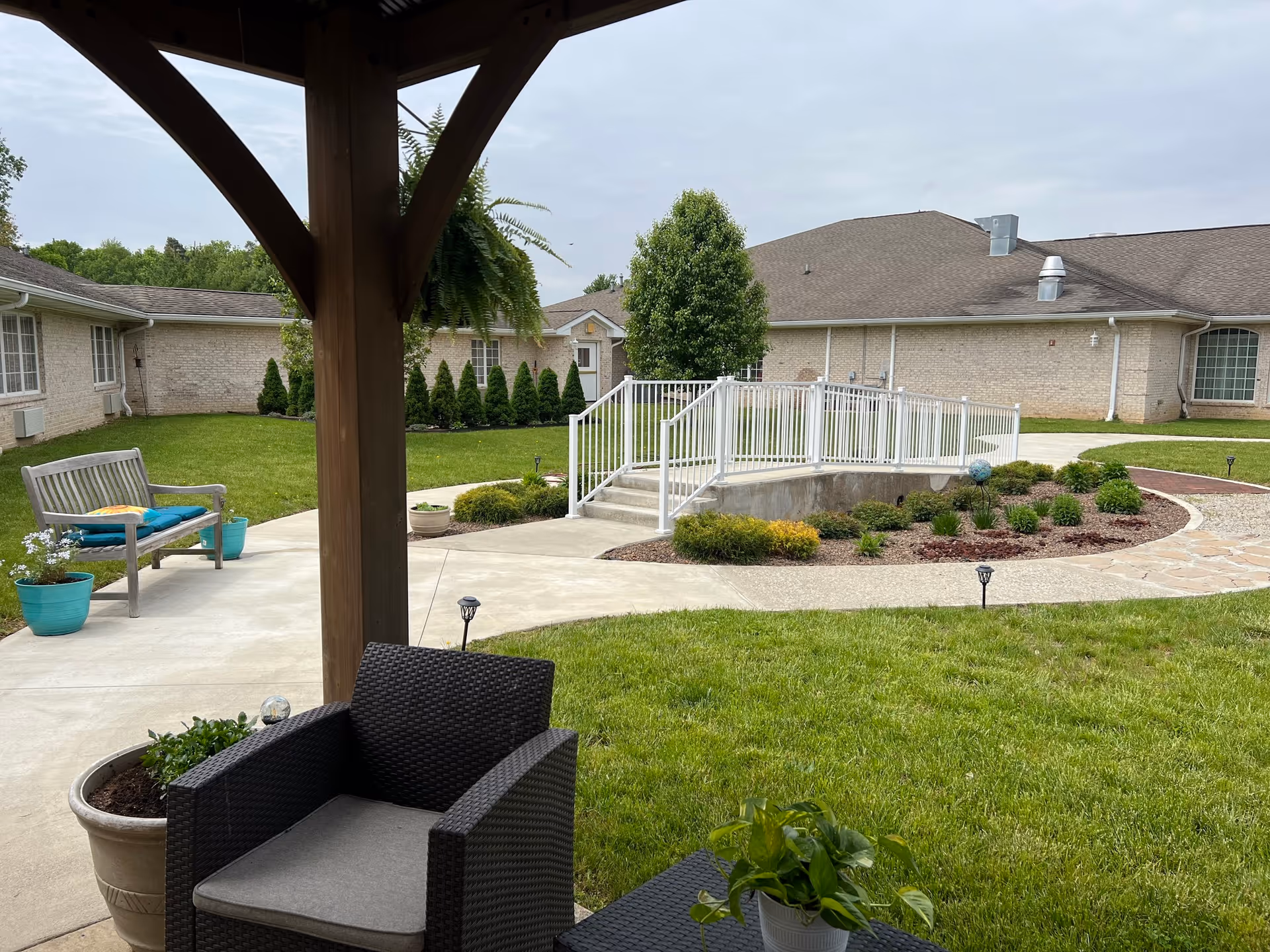 Outdoor patio area at Avon Health and Rehabilitation Center featuring a wooden pergola with hanging plants, a black wicker chair with a cushion, a wooden bench with colorful pillows, potted plants, a concrete walkway, landscaped garden beds, and a building with beige brick walls and a gray roof in the background.