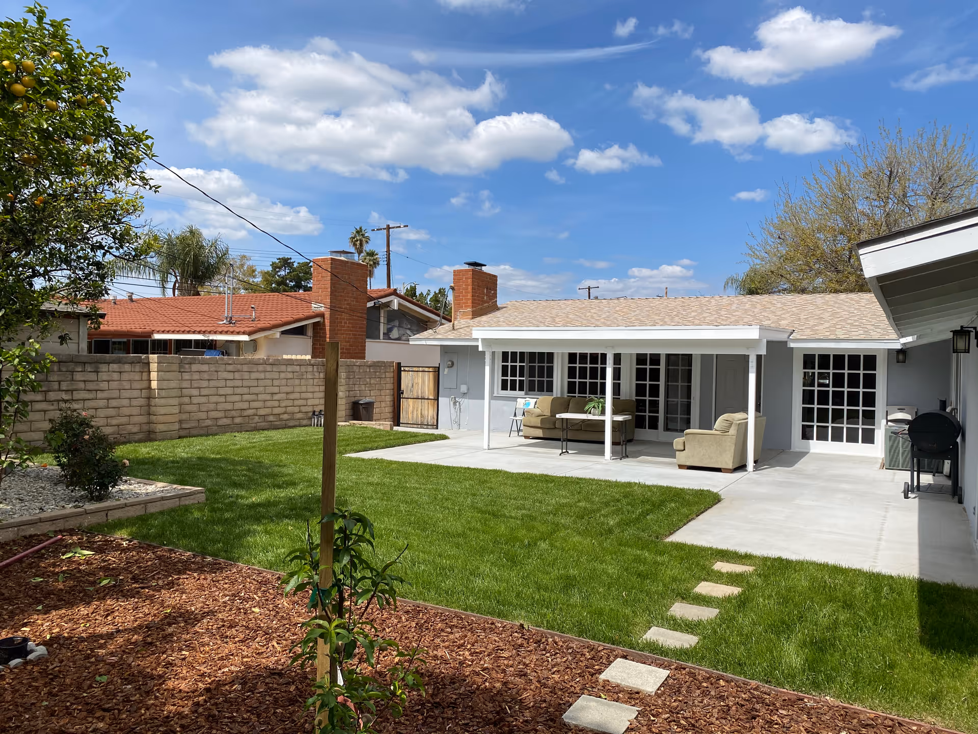 A backyard area of a senior care facility with a well-maintained green lawn, a small garden with mulch and plants, a concrete patio with outdoor seating including a sofa and armchair, and a barbecue grill. The building has light gray walls, multiple glass doors, and a beige shingled roof under a partly cloudy blue sky.