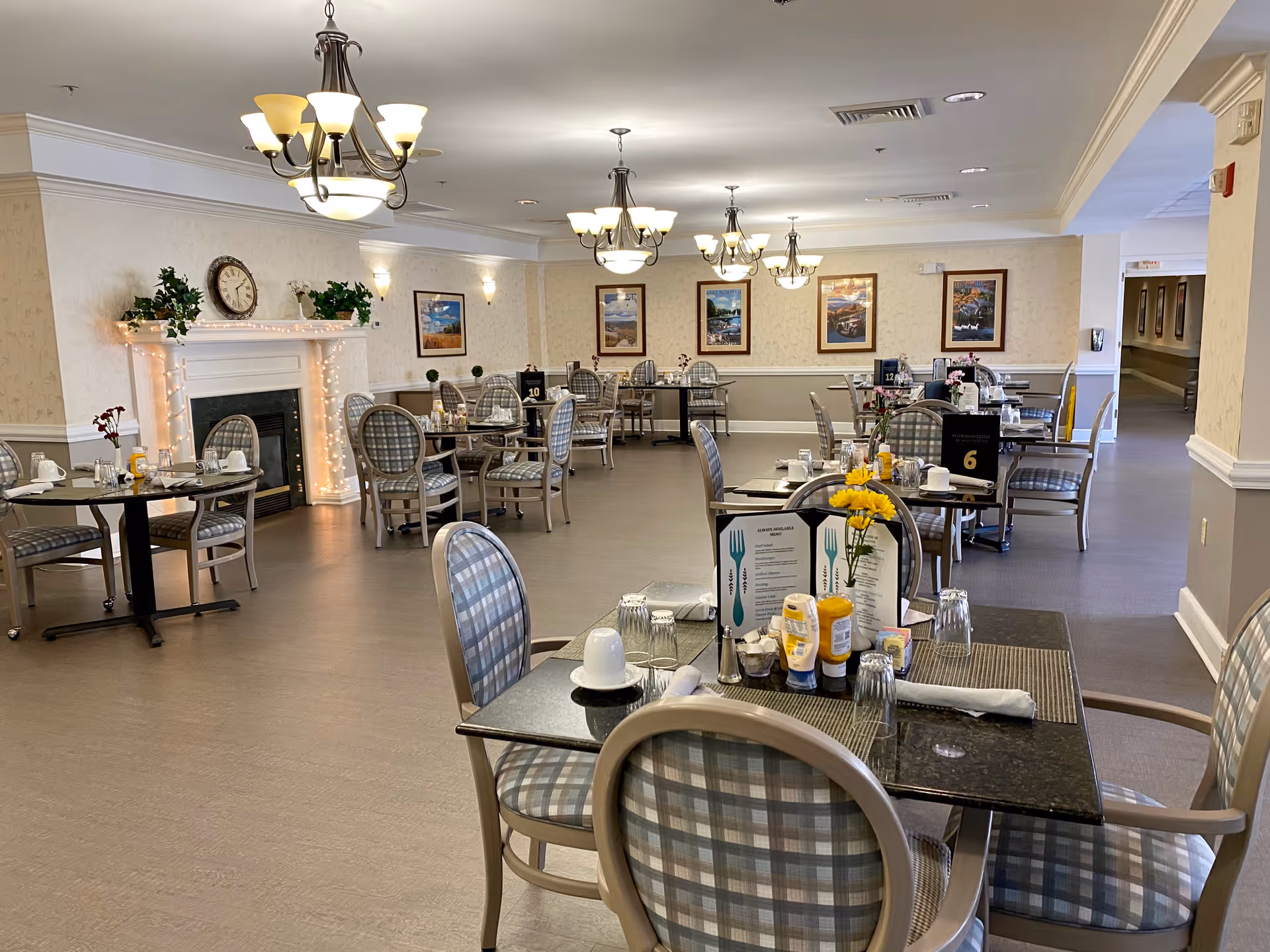 A spacious dining room with multiple tables and chairs arranged neatly. Each table is set with menus, glasses, cups, and condiments. The room features a decorative fireplace adorned with plants and a clock above it. Several framed pictures hang on the walls, and elegant chandeliers provide lighting from the ceiling.