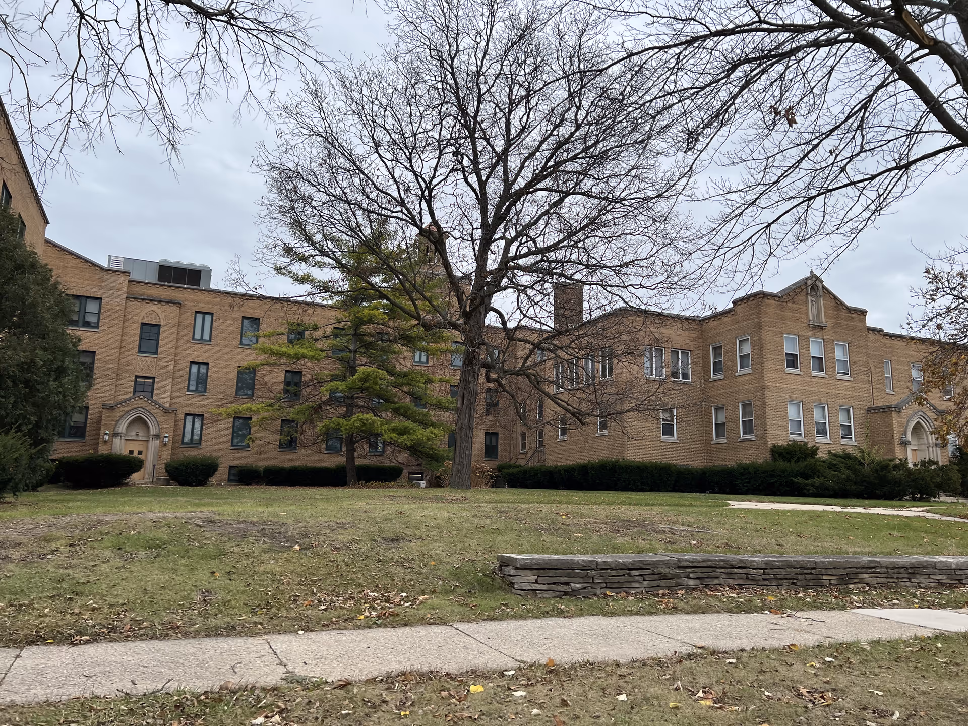 Exterior view of a large, multi-story brick building with multiple windows and two arched doorways. The building is surrounded by a grassy area with some bushes and leafless trees under an overcast sky.