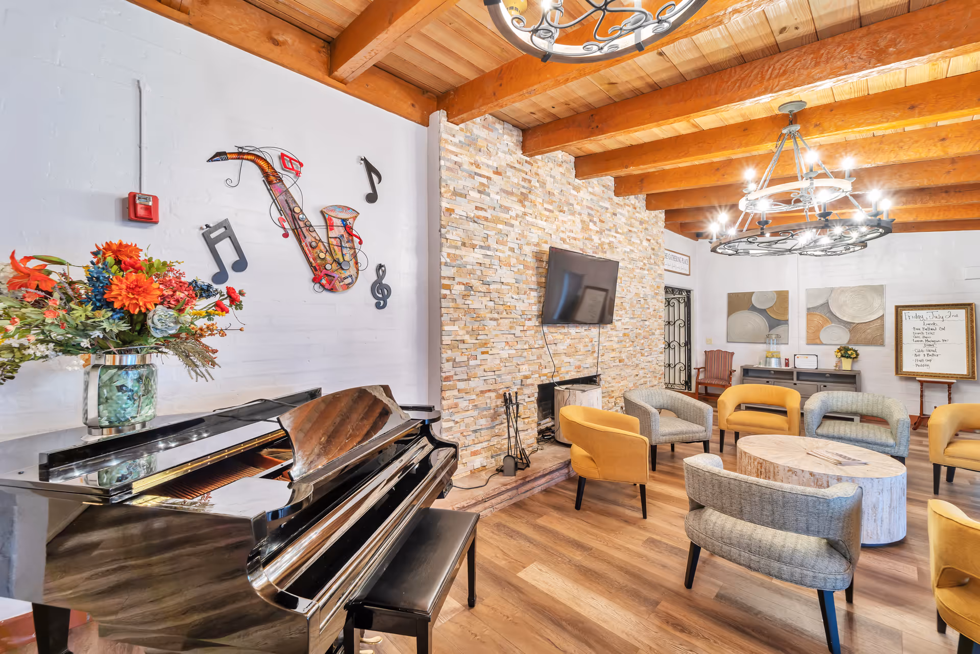 A cozy senior living common area with a black grand piano adorned with a vase of colorful flowers. The room features a stone fireplace with a flat-screen TV mounted above it, wooden ceiling beams, and modern chandeliers. Comfortable chairs in yellow and gray are arranged around a round wooden coffee table. Musical note decorations are mounted on the white brick wall above the piano.