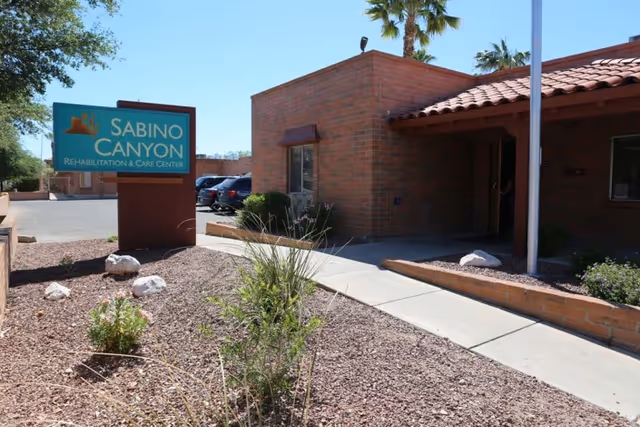 Exterior view of Sabino Canyon Rehabilitation & Care Center showing a brick building with a tiled roof, a sidewalk leading to the entrance, desert landscaping with rocks and plants, and a sign with the facility's name.