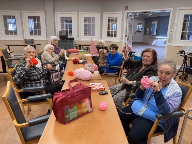 A group of elderly women sitting around a long wooden table in a well-lit room, engaging in knitting activities with yarn and knitting needles. The room has light-colored walls, several windows, and wooden flooring. Various balls of yarn and knitting supplies are spread on the table.