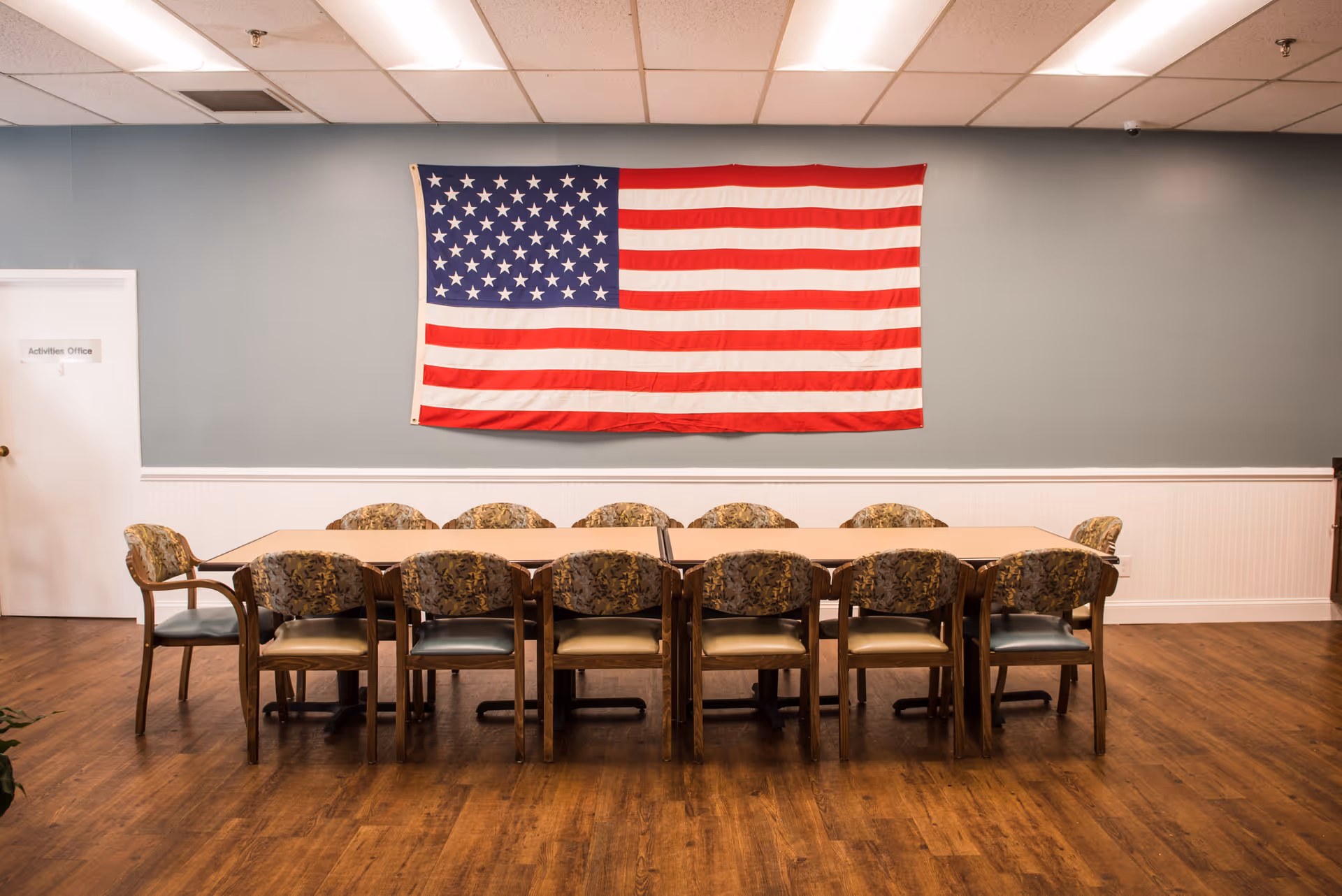 A meeting or activity room with a long table surrounded by twelve chairs with patterned upholstery. An American flag is hanging on the wall behind the table. There is a door labeled 'Activities Office' on the left side of the wall. The room has wooden flooring and a light blue wall with white wainscoting.