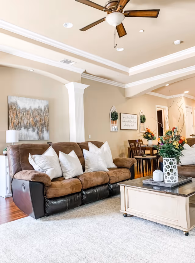 A cozy living room with a brown and black reclining sofa adorned with white pillows, a white wooden coffee table with a decorative vase of flowers, a ceiling fan with wooden blades, and a dining area with a wooden table and chairs in the background. The walls are beige with white trim and decorated with artwork and greenery.