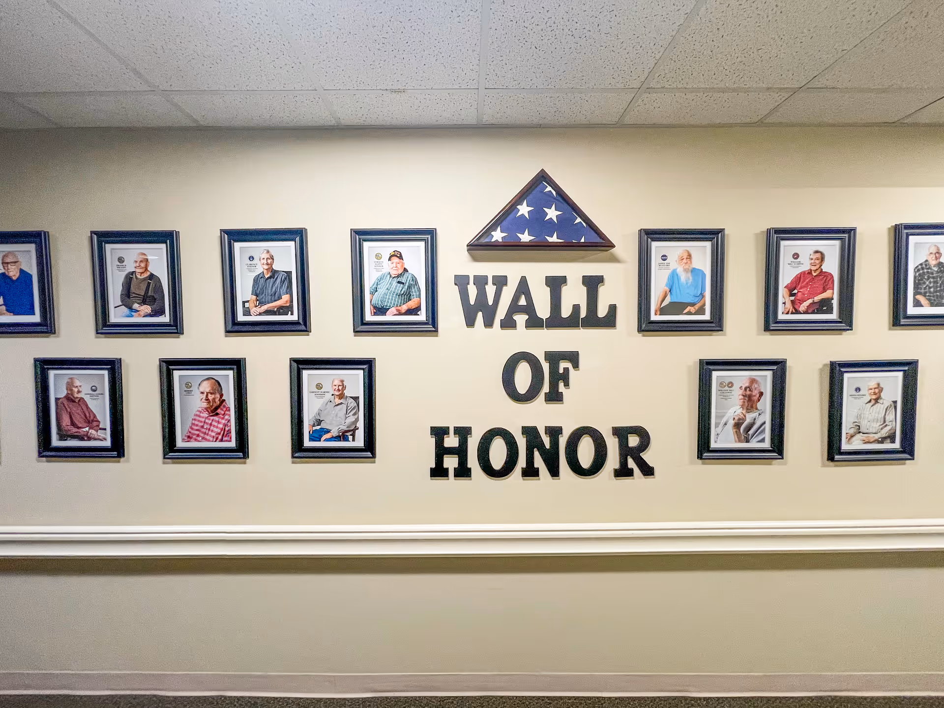 A wall display titled 'Wall of Honor' featuring framed portraits of elderly individuals arranged in two rows with a folded American flag in a triangular case centered above the text.
