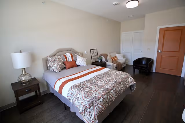 A neatly made bed with patterned bedding and multiple pillows in a senior living bedroom. Next to the bed is a dark wooden nightstand with a modern lamp. In the background, there is a small couch with cushions and a framed picture resting on the floor against the wall. The room has dark wooden flooring, a white closet door, and a closed orange door.