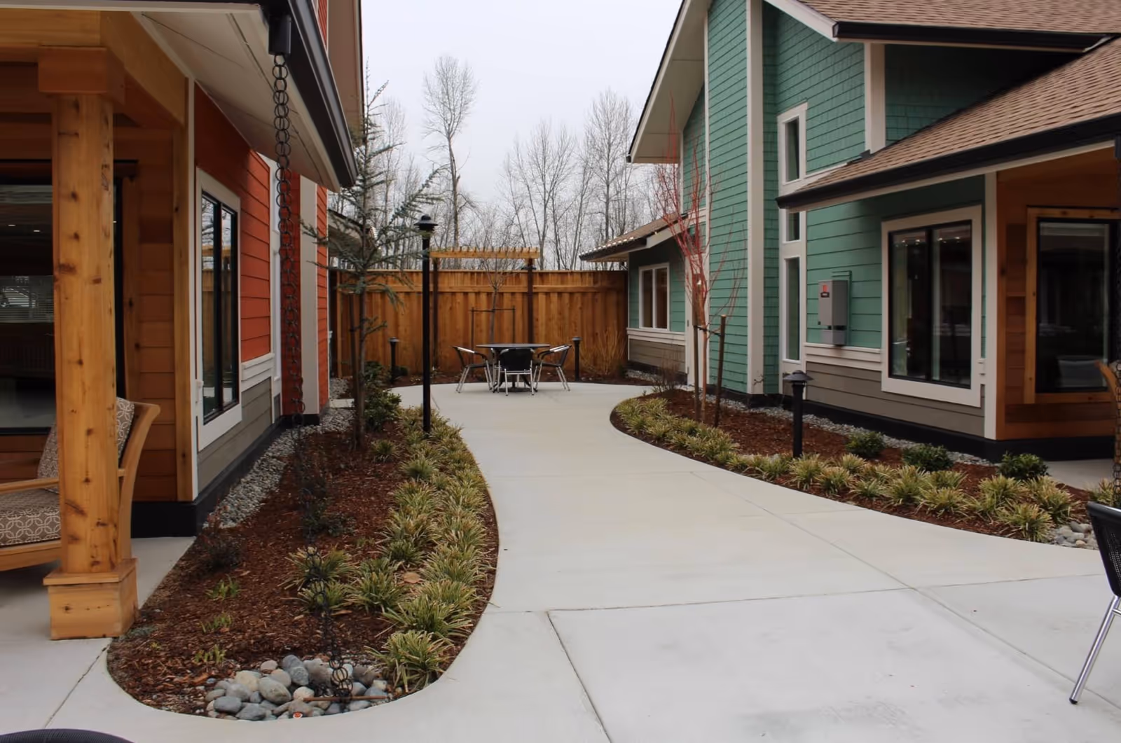 Outdoor courtyard area between two buildings with a concrete walkway, landscaped garden beds with plants and small trees, and a round table with four chairs at the end of the path. The buildings have wood and green siding with large windows.