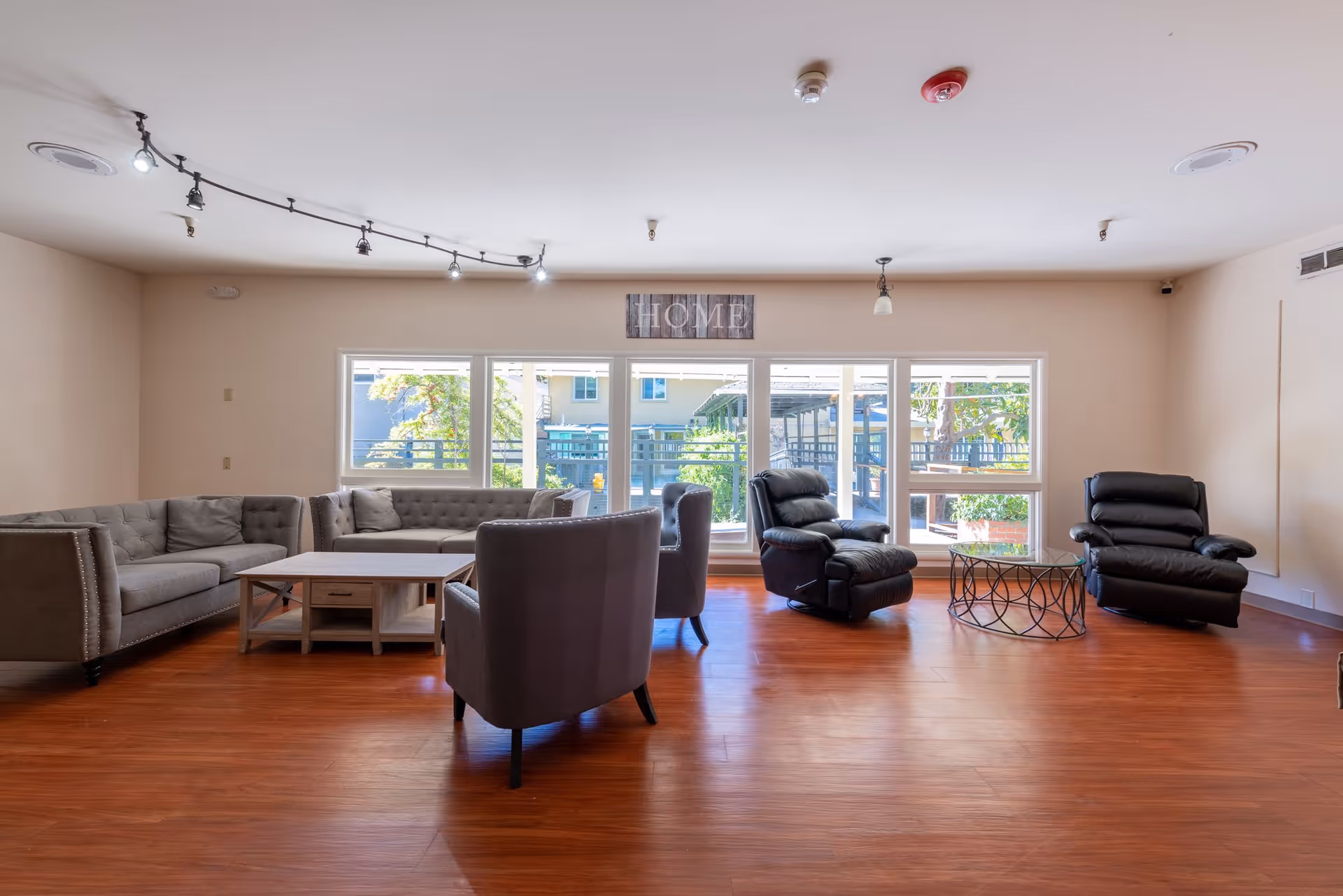 A spacious living room with wooden flooring featuring two gray sofas, two gray armchairs, and two black recliners arranged around two coffee tables. Large windows in the background allow natural light to fill the room, and a decorative sign reading 'HOME' is mounted above the windows.