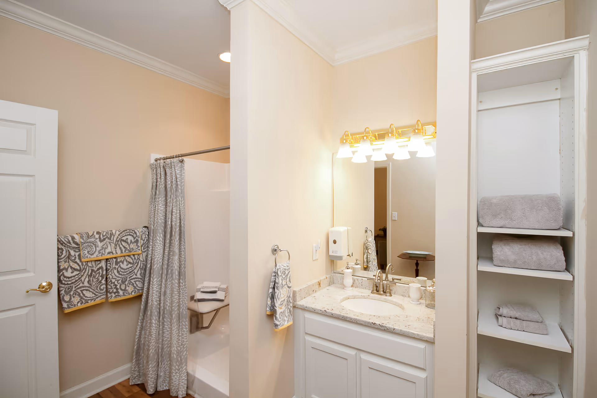 A clean bathroom with a white vanity and granite countertop, a large mirror with six lights above it, a towel ring with a patterned hand towel, a shower with a gray patterned curtain, and built-in shelves holding neatly folded gray towels.