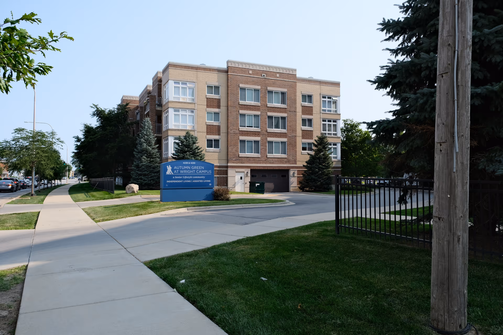 Exterior view of a multi-story senior living building with a sign in front that reads 'Autumn Green at Wright Campus, a Senior Lifestyle community, Independent Living | Assisted Living'. The building is surrounded by trees, grass, and a sidewalk leading up to the entrance.