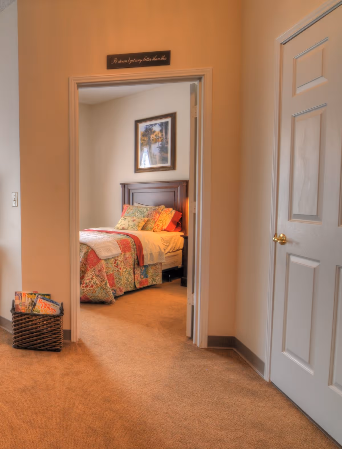 View through a doorway into a bedroom with a bed covered in a colorful patchwork quilt and multiple pillows. A framed picture hangs on the wall above the bed. A basket with magazines is placed on the carpeted floor outside the bedroom. The walls are painted beige and there is a closed white door with a gold handle on the right side of the image.