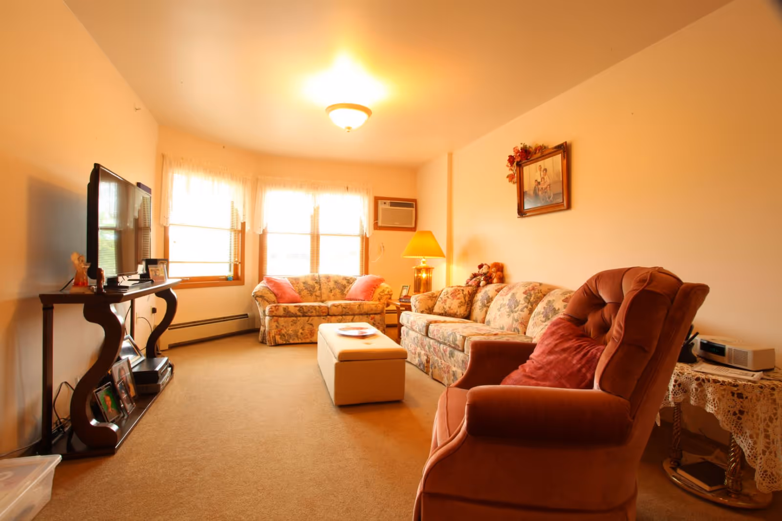 A cozy living room with beige carpet and cream-colored walls. The room features a floral-patterned sofa and loveseat, a rust-colored armchair with a matching pillow, a rectangular ottoman in the center, and a wooden TV stand holding a flat-screen television and framed photos. There is a table with a lace tablecloth and a clock radio on the right side. The room is softly lit by a ceiling light and a table lamp near the sofa. Three windows with sheer white curtains allow natural light to enter.