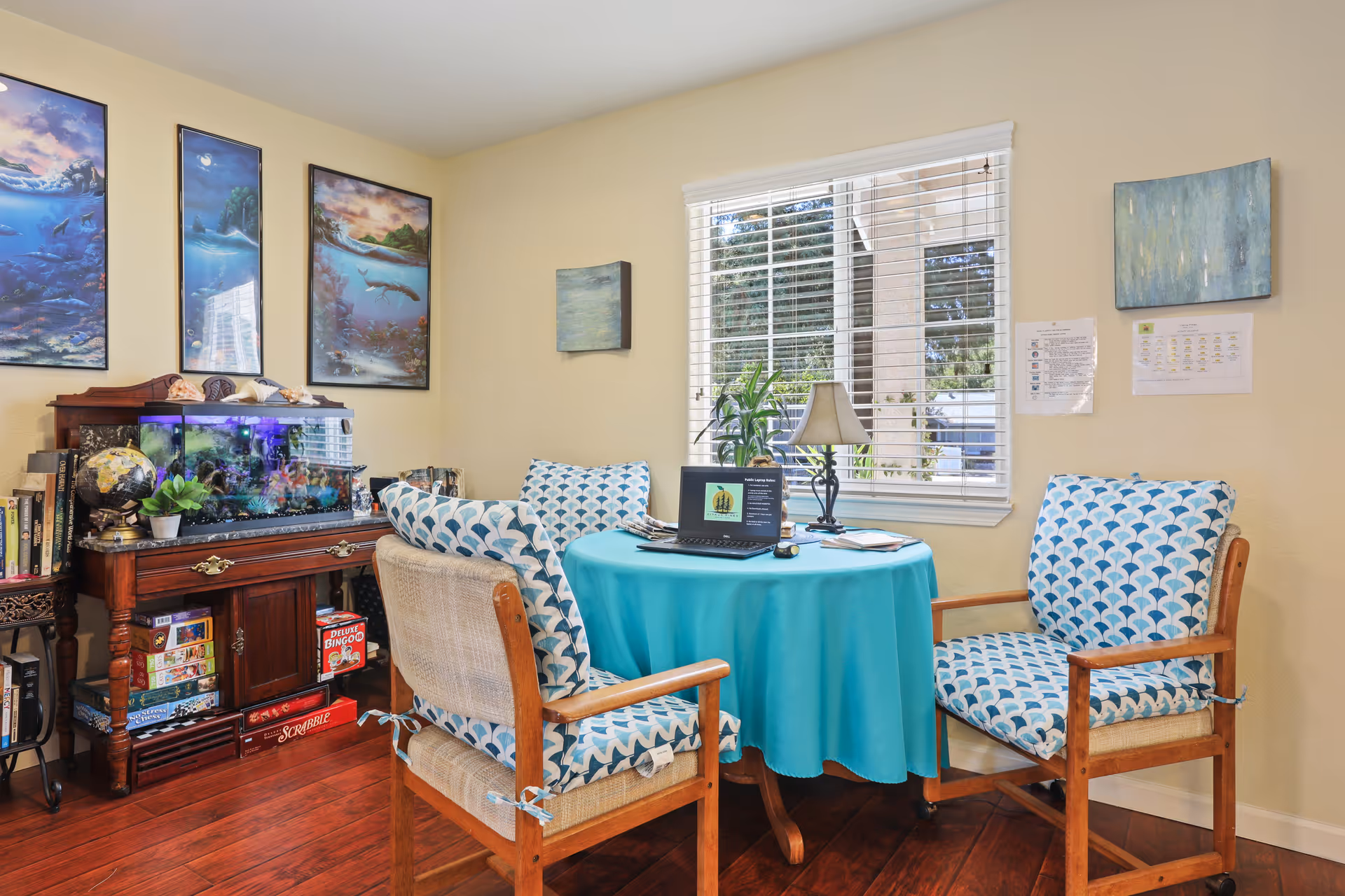A cozy sitting area in a senior living facility with a round table covered by a turquoise tablecloth, surrounded by four wooden chairs with blue and white patterned cushions. On the table is a laptop, a lamp, and some papers. Behind the table is a window with white blinds, and on the walls are several paintings. To the left is a wooden cabinet with an aquarium on top and various board games stored underneath.
