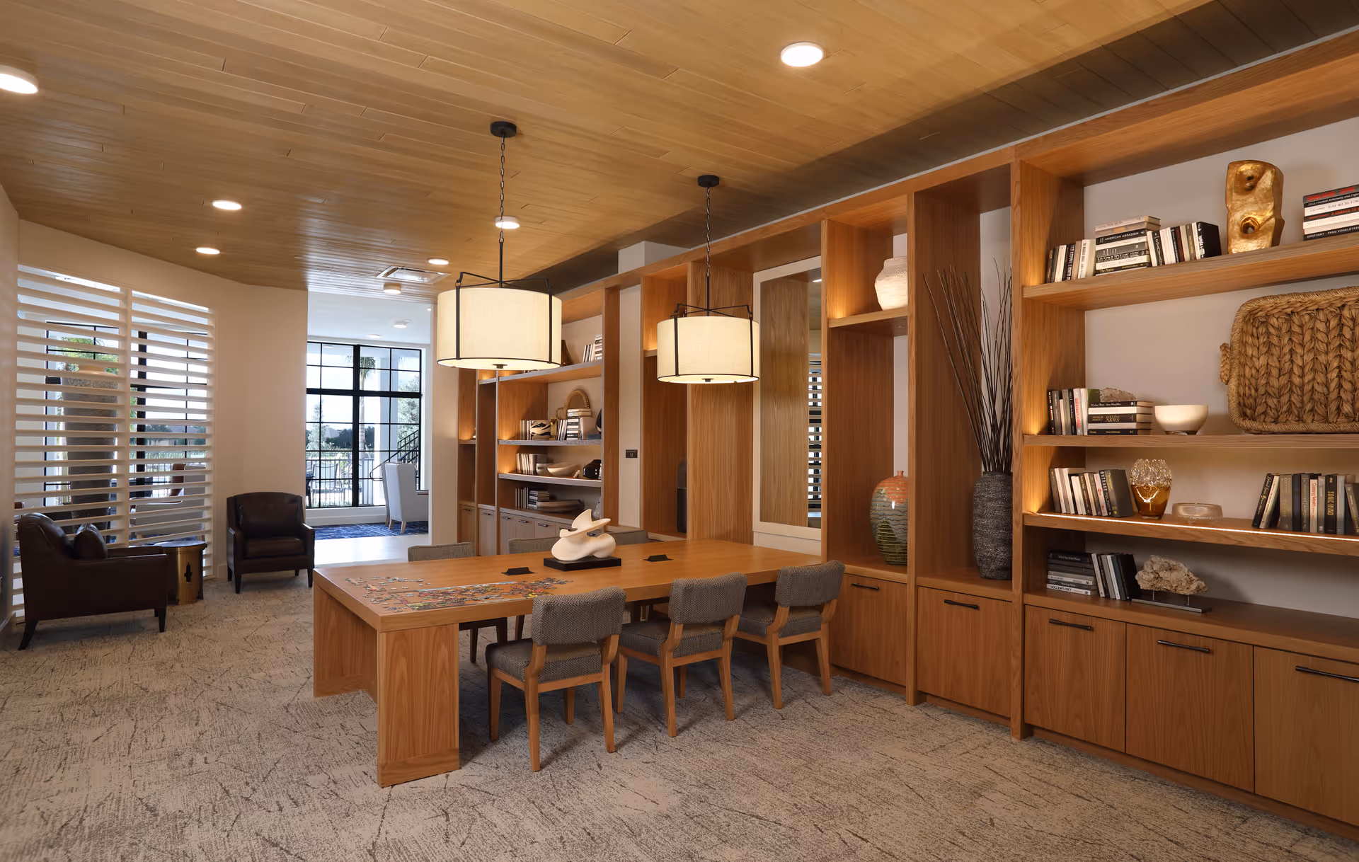 A modern, well-lit common area with a large wooden table surrounded by six chairs. The room features built-in wooden shelves filled with books and decorative items, two hanging pendant lights above the table, and a seating area with two leather armchairs near a window with horizontal blinds. The ceiling is wooden, and the floor is carpeted.