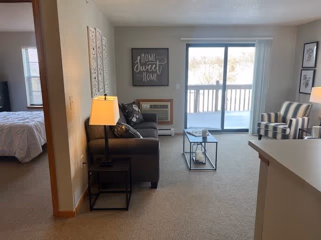 A cozy living room area with a gray sofa, a side table with a lamp, a coffee table, and a striped armchair. There is a sliding glass door leading to a balcony with a snowy outdoor view. On the wall above the sofa is a decorative sign that reads 'Home Sweet Home'. To the left, a bedroom with a bed and window is partially visible.