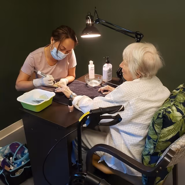 A caregiver wearing a face mask and gloves is giving a manicure to an elderly woman seated in a chair with a walker nearby. The setting appears to be a cozy indoor space with a dark green wall and a desk lamp illuminating the work area.