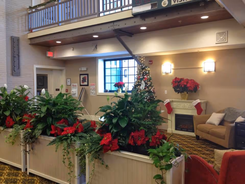 Interior view of a senior community common area decorated for Christmas with a Christmas tree, poinsettias, and stockings hanging on a fireplace mantel. There are comfortable chairs and a sofa, plants in planters, and a balcony railing above.