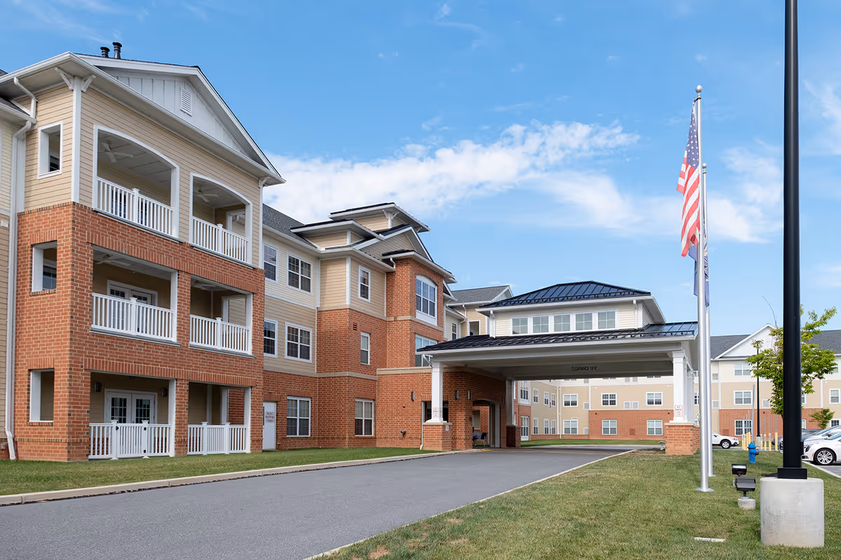 Exterior view of a multi-story senior living facility building with brick and beige siding, featuring balconies and a covered entrance driveway. An American flag and another flag are flying on flagpoles near the driveway under a clear blue sky.