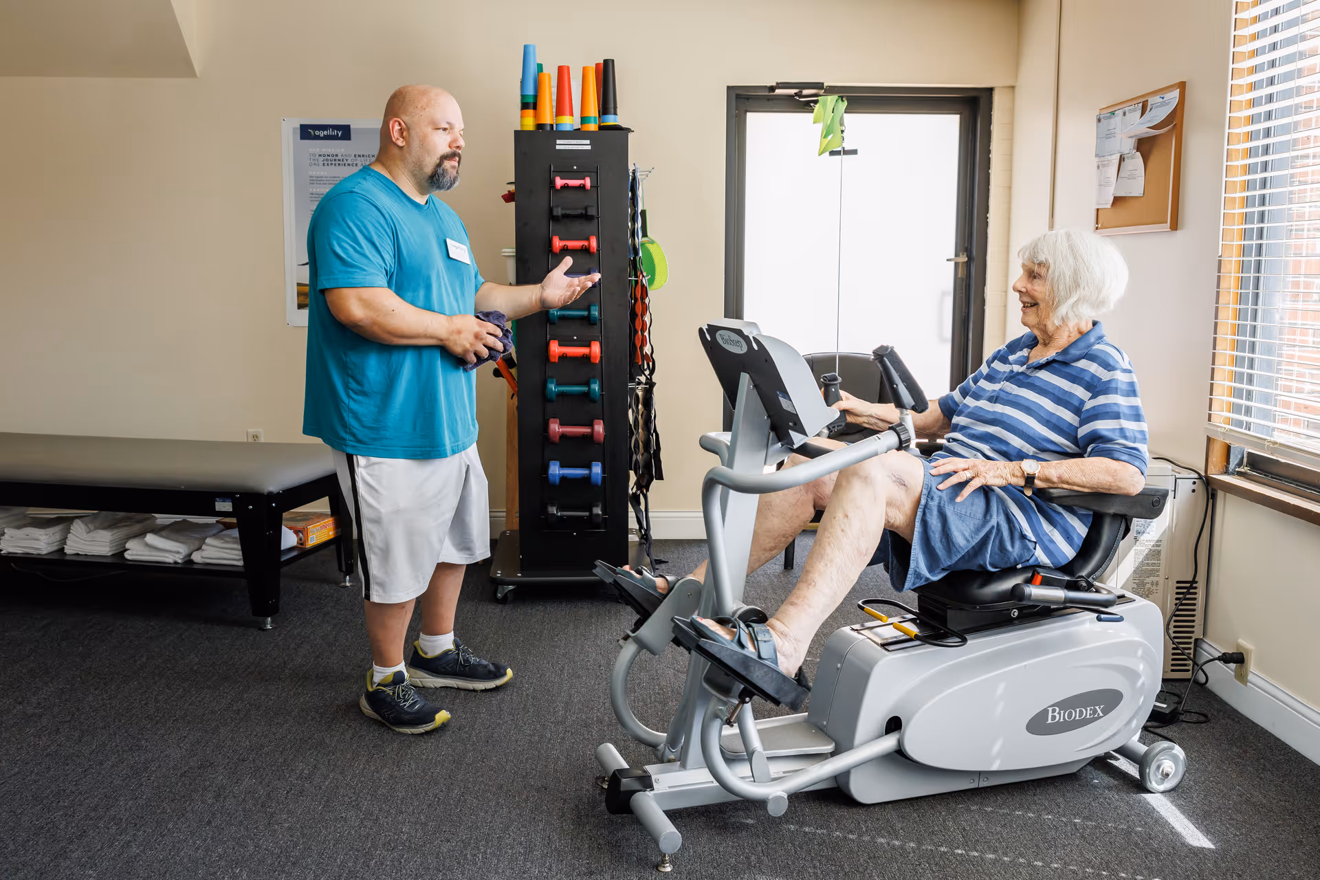 An elderly woman in a striped blue shirt and shorts is exercising on a recumbent stationary bike in a fitness room. A man in a teal shirt and white shorts stands nearby, talking to her. The room has exercise equipment, a bench with folded towels, and a window with blinds letting in natural light.