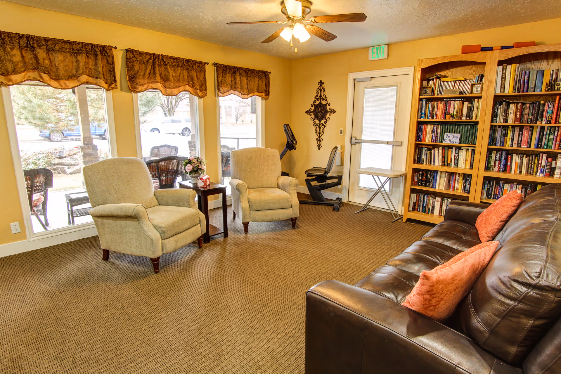 A cozy living room area with two beige armchairs and a small wooden side table between them, a dark brown leather sofa with two orange cushions, a large wooden bookshelf filled with books, a stationary exercise bike near a door with a window, and three large windows with brown valances letting in natural light.