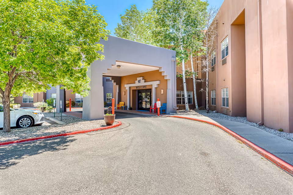 Entrance to a senior living facility with a covered drop-off area, a 'Welcome' sign above the door, colorful chairs near the entrance, surrounded by trees and a parked white car on the left side.